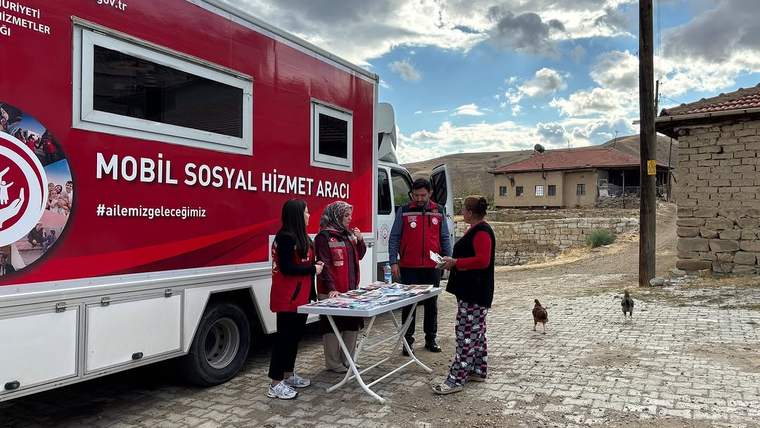 Kırmızı mobil sosyal hizmet aracı yanında üç kişi, broşürlerin bulunduğu masanın başında duruyor; arkada taş evler var.
