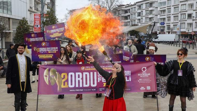 Meydanda mor pankart taşıyan kalabalık önünde, ateş gösterisi yapan kostümlü bir kişi görülüyor.