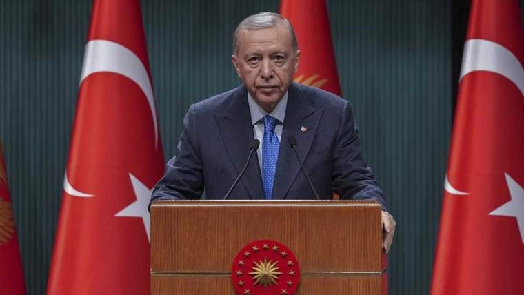 Kürsüde konuşan bir kişi, arkasında Türk bayraklarıyla birlikte görünüyor.
