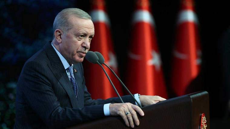Kürsüde mikrofonlara konuşan bir kişi, arka planda kırmızı bayraklarla görülüyor.