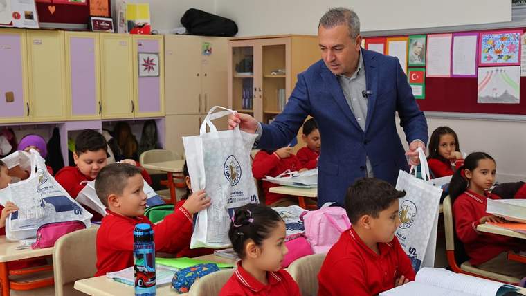 Sınıfta, kırmızı tişörtlü çocuklar masalarında oturuyor; bir kişi çocuklara kırtasiye yardımı dağıtıyor.