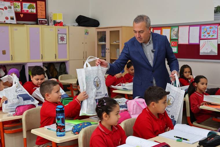Sınıfta, kırmızı tişörtlü çocuklar masalarında oturuyor; bir kişi çocuklara kırtasiye yardımı dağıtıyor.