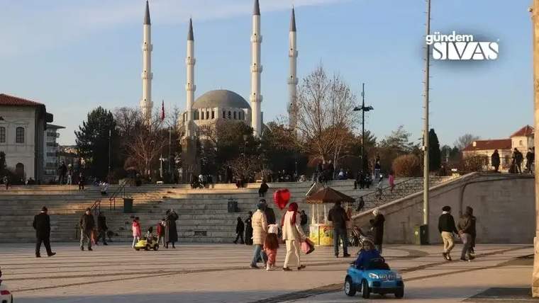 Güneşli havada, önünde insanların bulunduğu geniş meydanın arkasında kubbeli ve minareli bir cami görülüyor.
