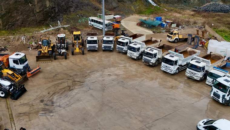 Kayalık yamaçların önündeki açık alanda, iş makineleri, kamyonlar ve servis araçları sıralanıyor.
