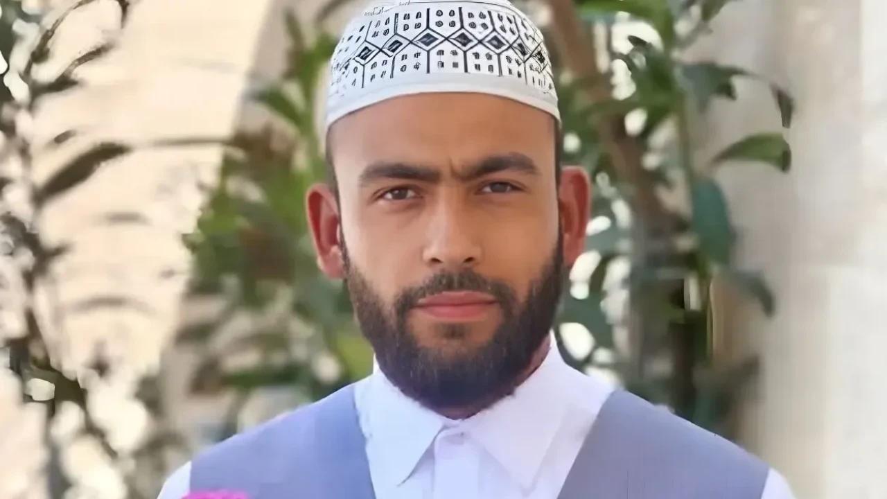 Beyaz desenli takke ve beyaz yakalı gömlek giyen sakallı bir kişi, bitkilerin önünde kameraya bakıyor.