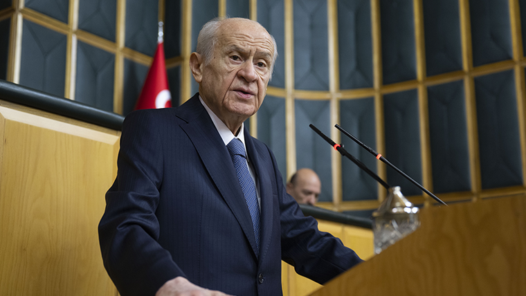 Kürsüde konuşan bir kişi, arka planda Türk bayrağı ve diğer kişiler görünüyor.