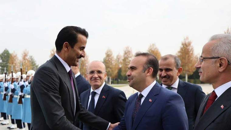 Takım elbiseli beş kişi açık havada tokalaşırken arkada üniformalı bir grup ve ağaçlar görülüyor.