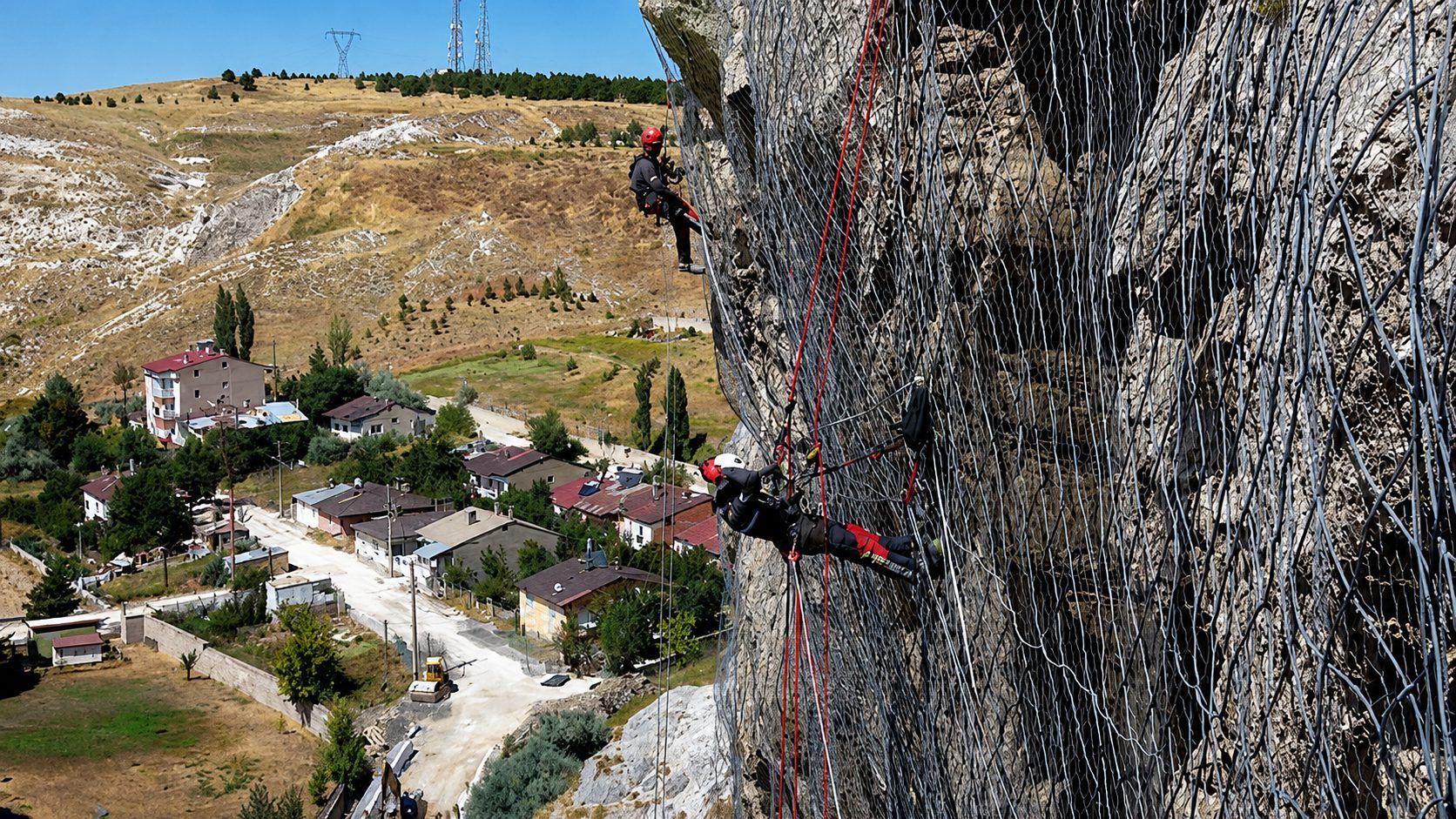 Kayalık yamaçta halatlarla çalışan iki kişi, aşağıdaki evlerin ve çevredeki arazinin üzerinde görülüyor.