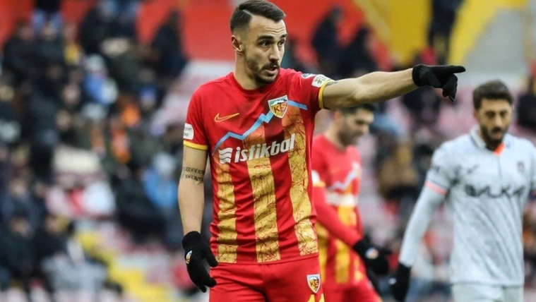 Kırmızı ve sarı renkte forma giymiş bir futbolcu, sahada işaret ederek bir yere bakıyor.