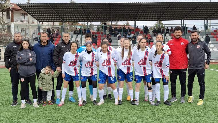 Futbol sahasında forma giyen genç kadın oyuncular, teknik ekip ve çocuklarla birlikte yan yana duruyor.