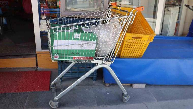 Market girişinin önünde, içinde yeşil kasa bulunan alışveriş arabası ve turuncu sepet duruyor.