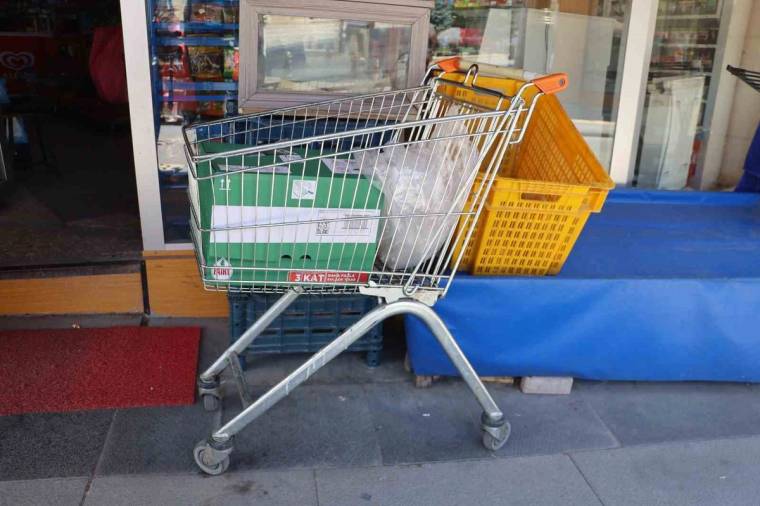 Market girişinin önünde, içinde yeşil kasa bulunan alışveriş arabası ve turuncu sepet duruyor.