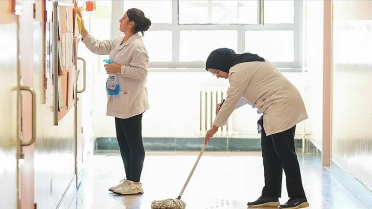 Koridorda iki kişi temizlik yapıyor; biri zemini paspaslarken diğeri duvardaki yüzeyi siliyor.