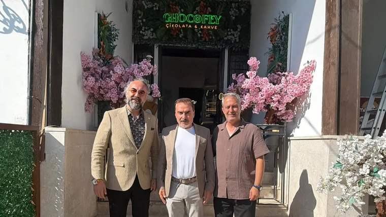 Üç kişi, bir kafenin önünde poz veriyor; arka planda çiçeklerle süslenmiş bir giriş var.