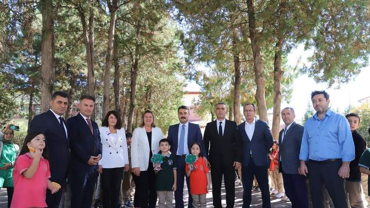 Ağaçlarla çevrili alanda, çocuklar ve yetişkinlerden oluşan bir grup birlikte ayakta duruyor.