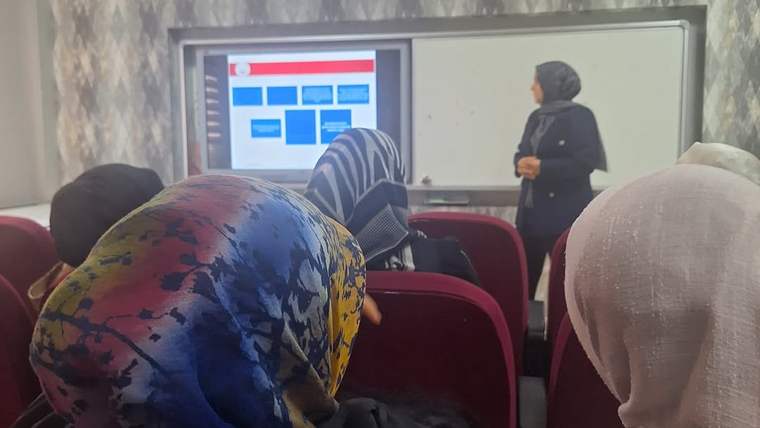 Bir sınıfta başörtülü kadınlar otururken, ön tarafta bir kadın bilgisayar ekranı ve beyaz tahta yanında duruyor.