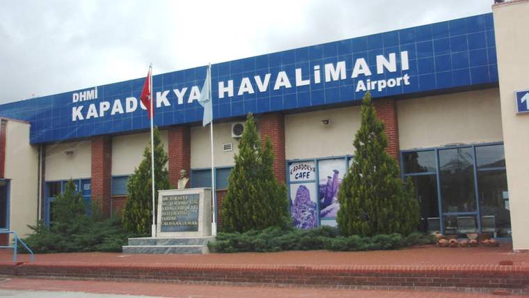Kapadokya Havalimanı binasının girişinde bayraklar, çam ağaçları ve kaldırım boyunca uzanan tabela görülüyor.