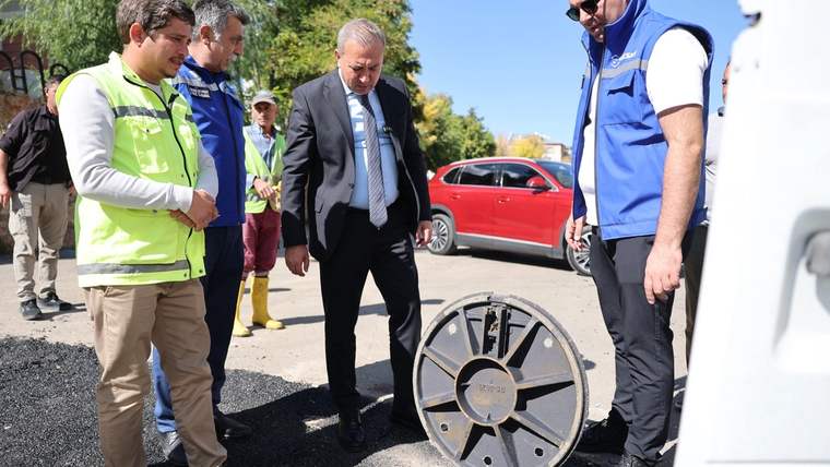 Yol kenarında üç kişi asfalt üzerindeki büyük metal kapağı incelerken, arkada kırmızı otomobil görülüyor.