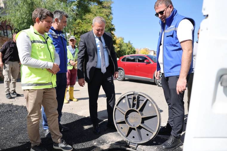 Yol kenarında üç kişi asfalt üzerindeki büyük metal kapağı incelerken, arkada kırmızı otomobil görülüyor.