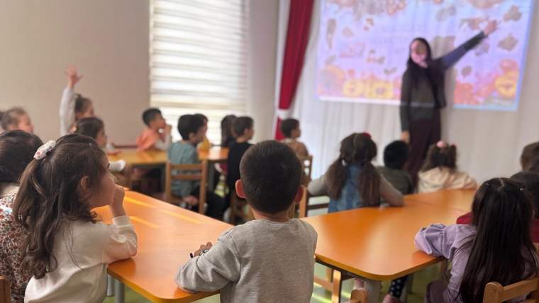 Sınıfta oturan çocuklar, duvardaki renkli sunumu gösteren bir kadın konuşmacıyı izliyor.