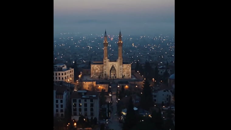 Şehir manzarasında, akşam saatlerinde aydınlatılmış bir cami ve çevresindeki binalar görünmekte.
