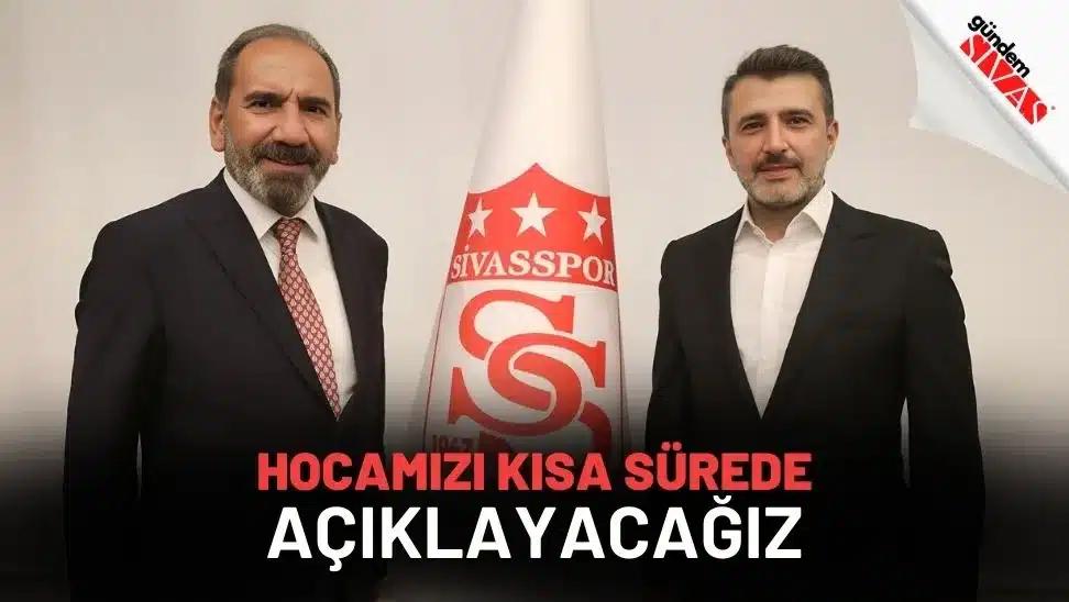 İki takım elbiseli kişi, üzerinde Sivasspor arması bulunan bayrağın önünde yan yana duruyor.