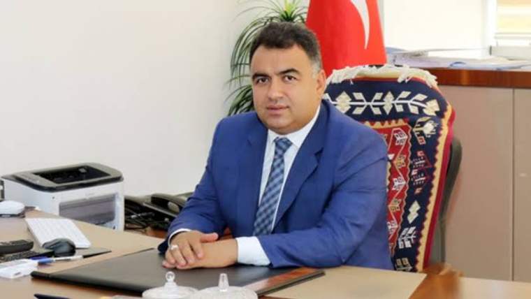 Mavi takım elbise giymiş bir kişi, masanın arkasında oturuyor ve önünde bir dosya var.