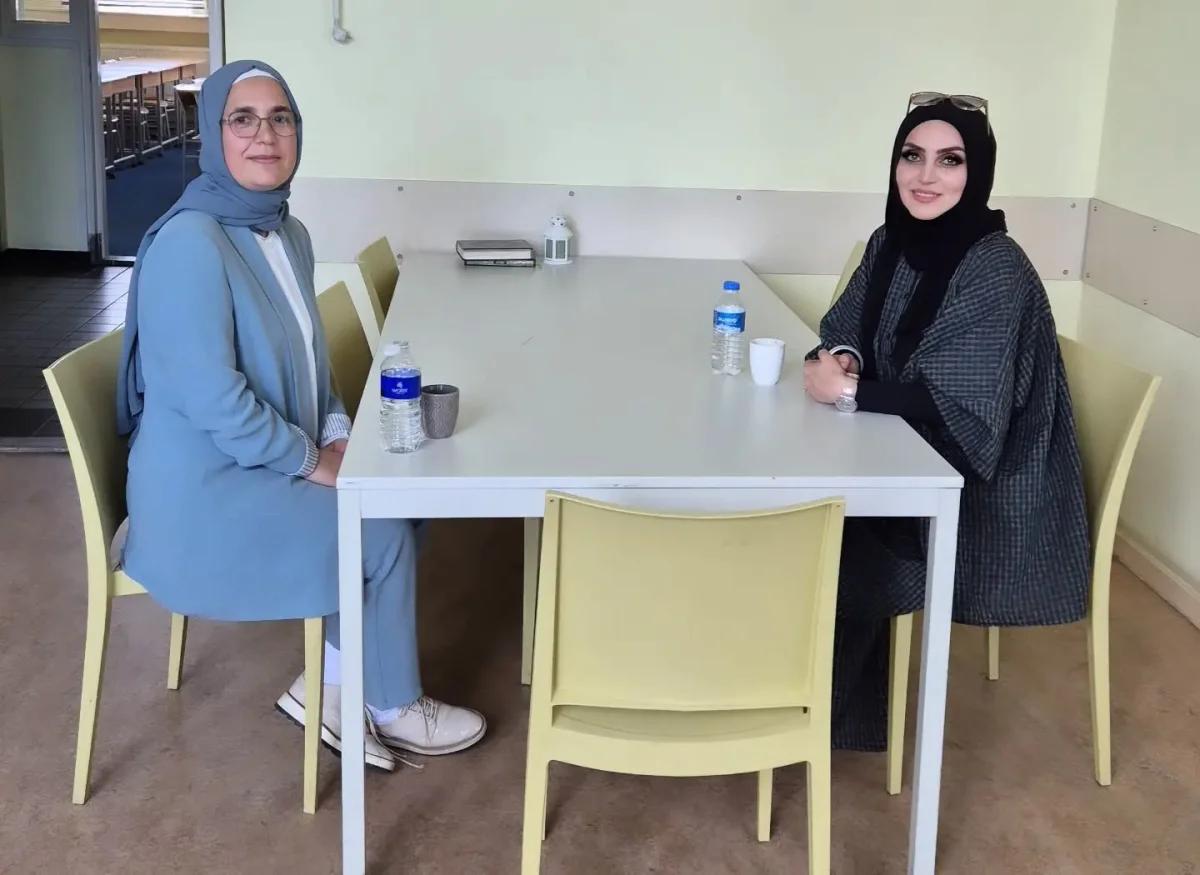 İki kadın, beyaz bir masanın etrafında oturuyor; masada su şişeleri ve bir fincan var.
