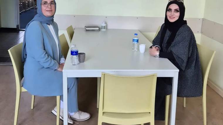 İki kadın, beyaz bir masanın etrafında oturuyor; masada su şişeleri ve bir fincan var.