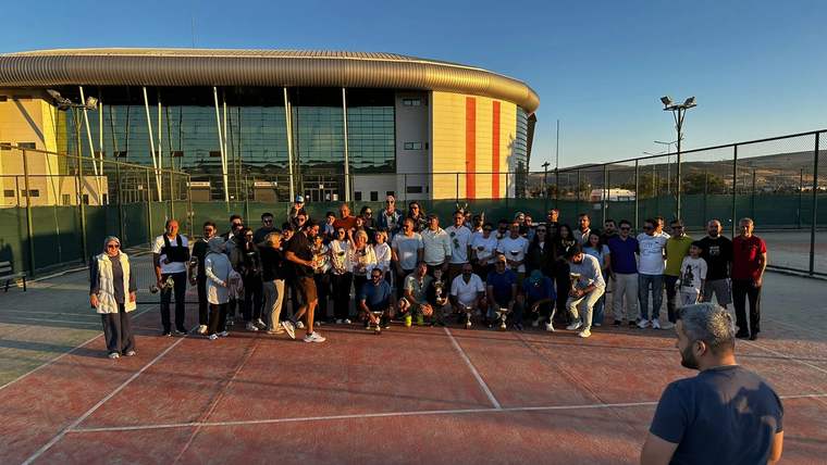 Tenis kortunda bir grup insan, arkalarında modern bir spor tesisi binasıyla birlikte poz veriyor.
