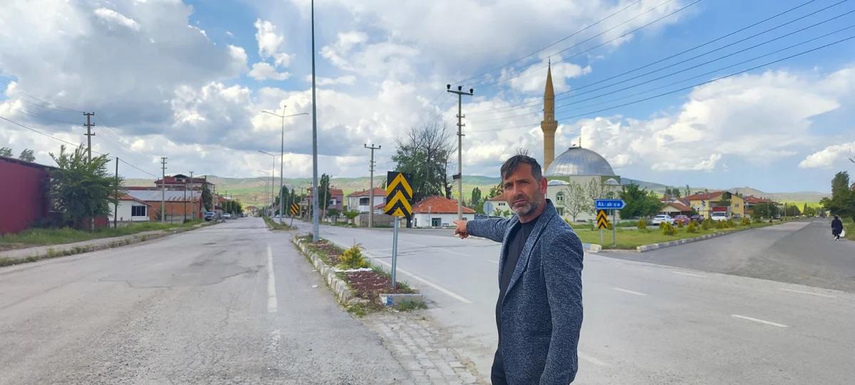 Bir adam, işaret ettiği yolda bulunan uyarı levhalarının önünde duruyor. Arkada bir cami görünmekte.