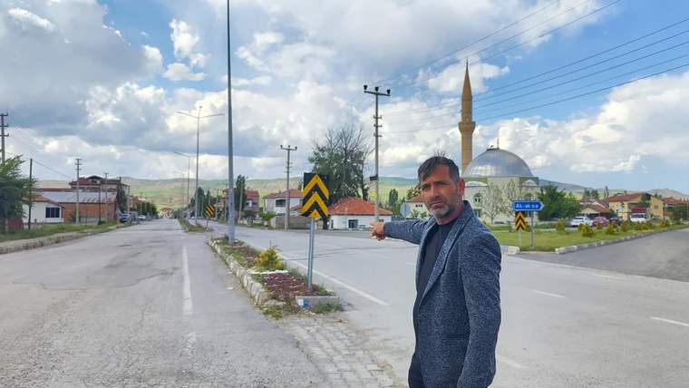 Bir adam, işaret ettiği yolda bulunan uyarı levhalarının önünde duruyor. Arkada bir cami görünmekte.
