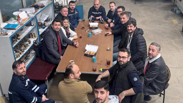 Atölye ortamında, uzun bir masa çevresinde oturan ve kameraya bakan bir grup erkek görülüyor.