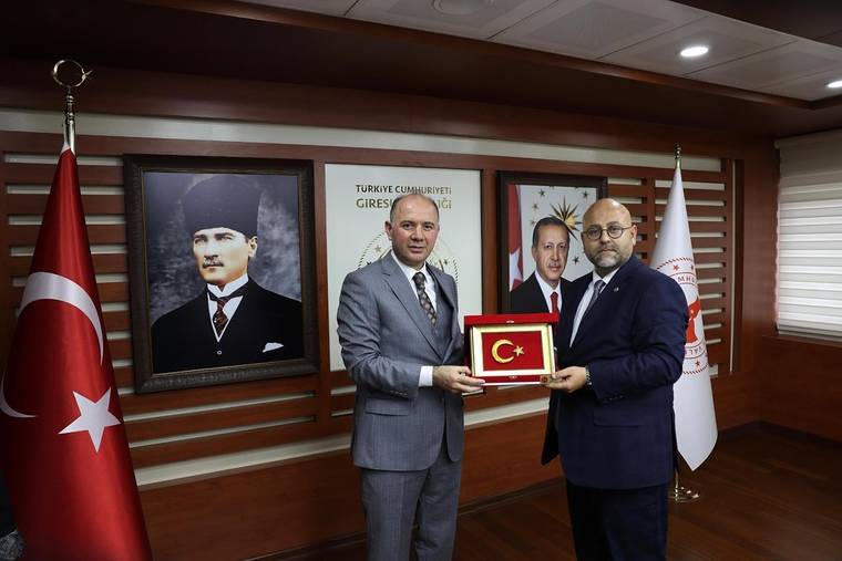 İki erkek, Türk bayrağı ve portrelerin bulunduğu ahşap kaplı odada plaket tutuyor.