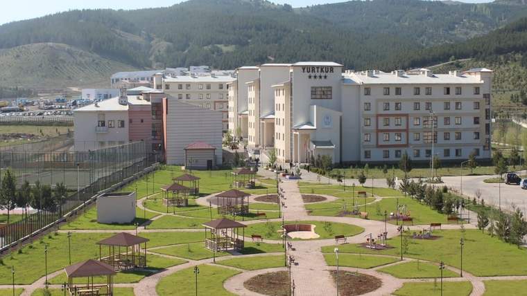 Yeşil alanlar ve yürüyüş yolları ile çevrili bir kampüs binası, arka planda dağlar görünmekte.