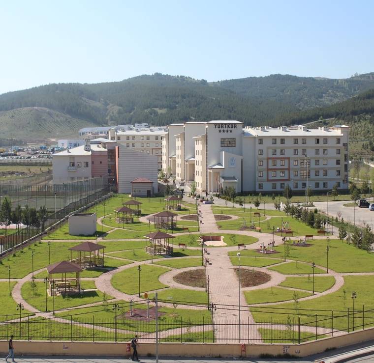 Yeşil alanlar ve yürüyüş yolları ile çevrili bir kampüs binası, arka planda dağlar görünmekte.