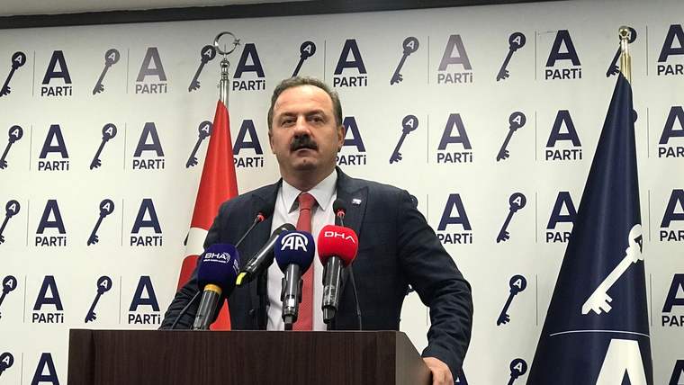 A PARTİ logolu kürsüde konuşan bıyıklı bir erkek, önündeki mikrofonların arasında duruyor; iki bayrak görülüyor.