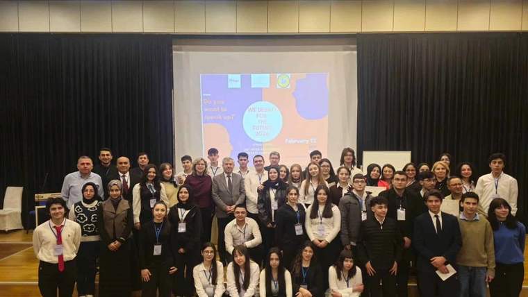 Bir grup öğrenci ve öğretmen, bir etkinlikte topluca poz veriyor. Arka planda büyük bir ekran var.