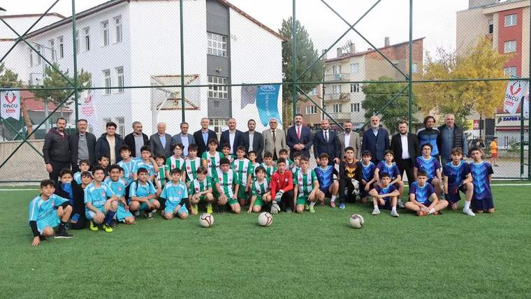 Çitlerle çevrili futbol sahasında farklı renklerde formalar giyen çocuklar ve yetişkinler, toplarla birlikte poz veriyor.