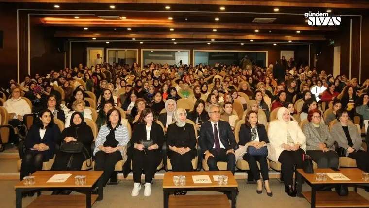 Bir etkinlikte oturan kalabalık bir kadın grubu, sahneye doğru bakıyor.