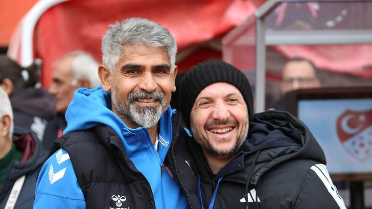 Mavi montlu, gri saçlı bir adam ile siyah bere ve mont giyen bir adam tribünlerde yan yana gülümsüyor.