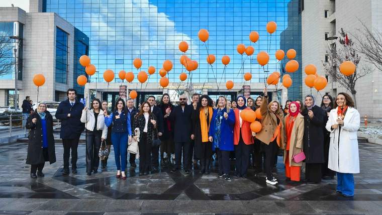 Kadınlar, elinde turuncu balonlar tutarak bir araya gelmiş, modern bir bina önünde duruyor.
