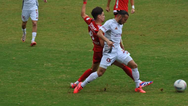 Futbolcular, yeşil sahada mücadele ediyor; biri beyaz, diğeri kırmızı forma giymekte.