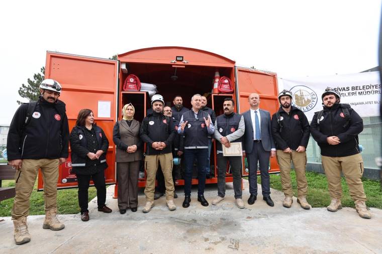 Turuncu bir aracın önünde, aralarında üniformalı kişilerin de bulunduğu bir grup yan yana duruyor.