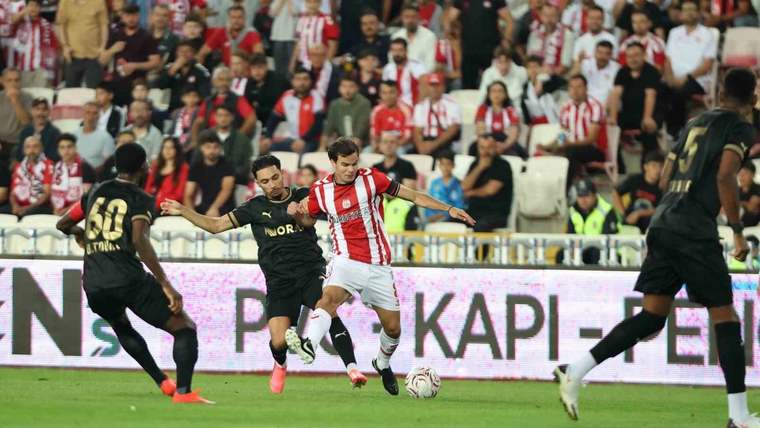 Futbol sahasında kırmızı-beyaz formalı oyuncu, rakip futbolcuların arasında topla ilerliyor; tribünler dolu.