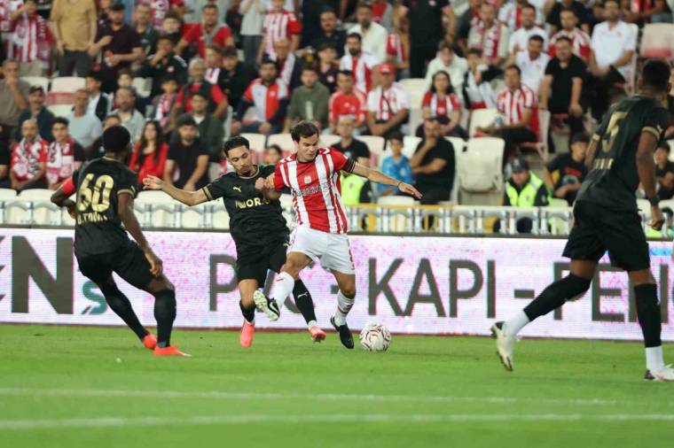 Futbol sahasında kırmızı-beyaz formalı oyuncu, rakip futbolcuların arasında topla ilerliyor; tribünler dolu.