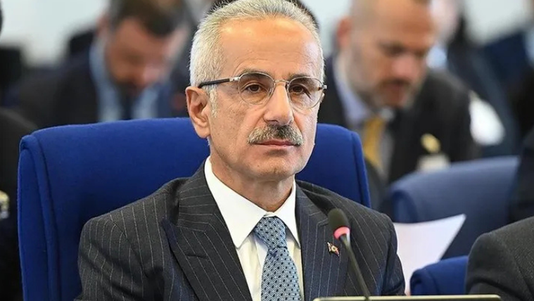 Mavi bir koltukta oturan, gözlük takan bir adam, dikkatle dinliyor.
