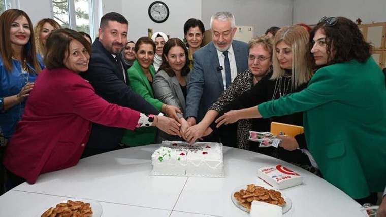 Bir grup insan, beyaz bir pasta keserken bir araya gelmiş. Masada çeşitli ikramlar var.