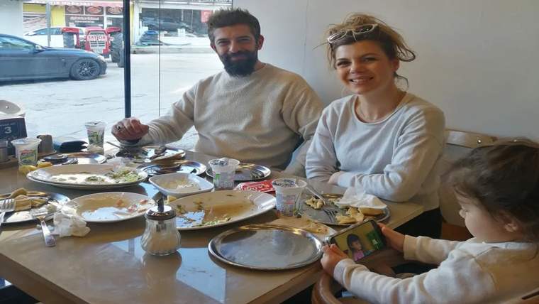Bir restoranda masada oturan sakallı bir erkek ve bir kadın, tabaklarla dolu sofrada gülümsüyor.