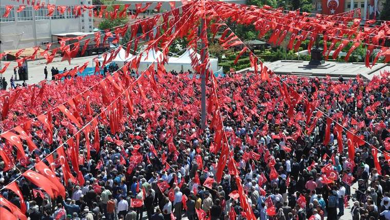 Türk bayrakları ve kırmızı flamalarla donatılmış alanda büyük bir kalabalık görülüyor.